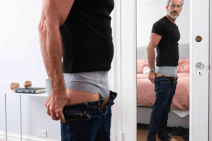 Man wearing gray absorbent underwear, looking in the mirror and pulling up his slacks.
