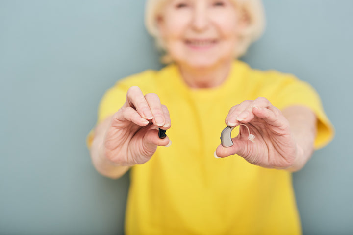 Older woman in a yellow shirt holding up hearing aids in her hands