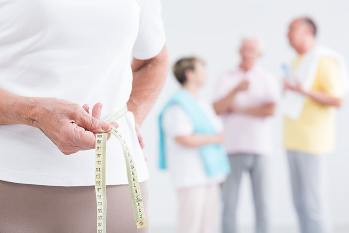 Man holding measuring tape around his waist to take measurements while other older adults are socializing in the background